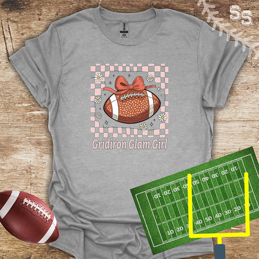 Gray t-shirt with 'Gridiron Glam Girl' design featuring a football and bow, on a wooden surface with a football and field graphic.